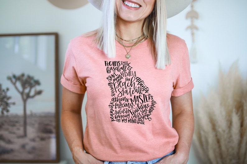 May include: A peach-colored t-shirt featuring a black graphic of the state of Georgia filled with the names of cities and phrases. The shirt has rolled-up sleeves and a crew neck.
