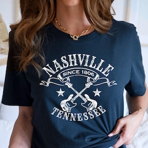 May include: Navy blue t-shirt with a white graphic featuring the text "NASHVILLE SINCE 1806 TENNESSEE" and crossed guitars. The design includes stars and a banner. A casual, music-themed tee.