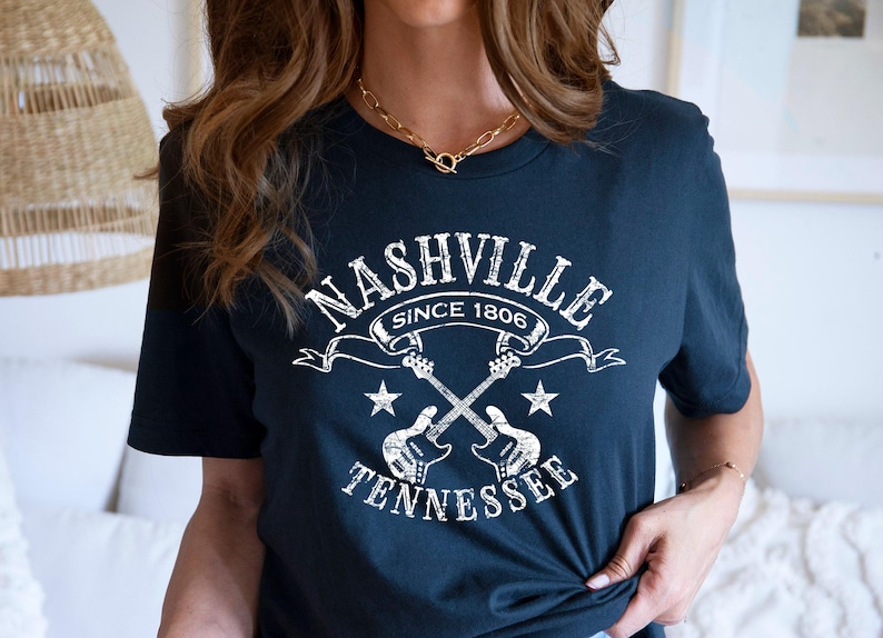 May include: Navy blue t-shirt with a white graphic featuring the text "NASHVILLE SINCE 1806 TENNESSEE" and crossed guitars. The design includes stars and a banner. A casual, music-themed tee.