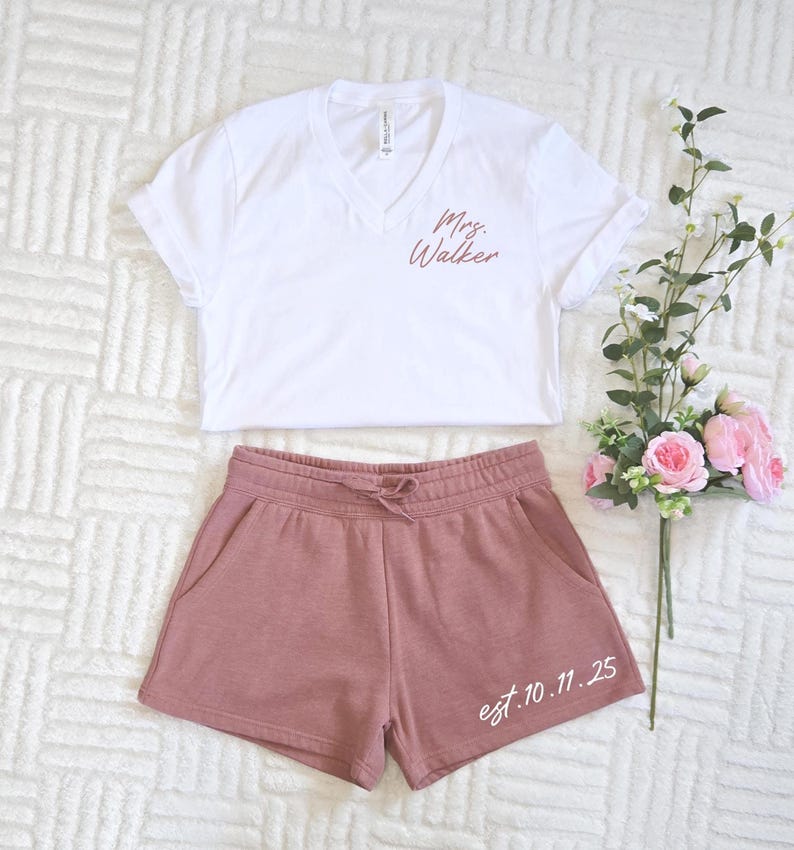 Op de afbeelding: Een witte V-hals T-shirt met "Mrs. Walker" in koperkleurige print, gecombineerd met mauve shorts. De shorts hebben een taille met trekkoord en de datum "est. 10.11.25" op de broekspijp. Een boeket roze rozen en groen ligt naast de kleding.