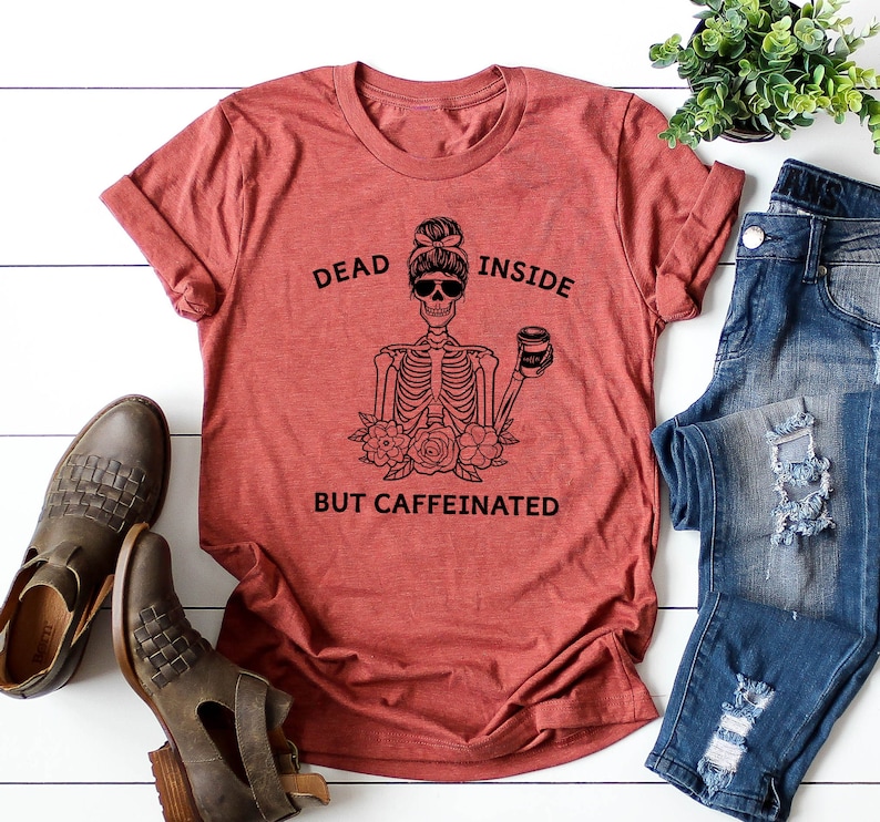 May include: Rust-colored t-shirt with the text "DEAD INSIDE BUT CAFFEINATED" and a skeleton graphic wearing sunglasses and holding a coffee cup. The graphic includes flowers. The shirt is paired with blue jeans and brown boots.