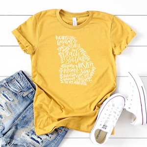 May include: Mustard yellow t-shirt with a white graphic in the shape of the state of Georgia, filled with text including city names and phrases. The shirt is paired with blue denim shorts and white sneakers.