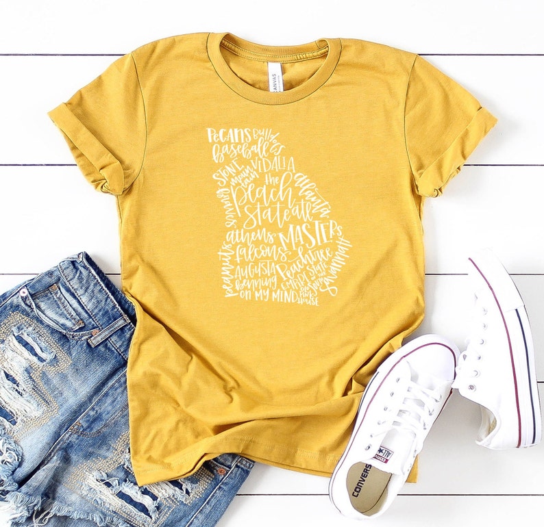 May include: Mustard yellow t-shirt with a white graphic in the shape of the state of Georgia, filled with text including city names and phrases. The shirt is paired with blue denim shorts and white sneakers.