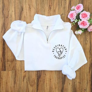 May include: White quarter-zip sweatshirt with the words "Respiratory Therapist" encircling a heart-shaped design with a stethoscope and EKG line. The sweatshirt has a high collar and long sleeves. Pink roses are in the background.