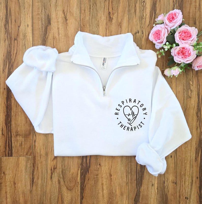 May include: White quarter-zip sweatshirt with the words "Respiratory Therapist" encircling a heart-shaped design with a stethoscope and EKG line. The sweatshirt has a high collar and long sleeves. Pink roses are in the background.