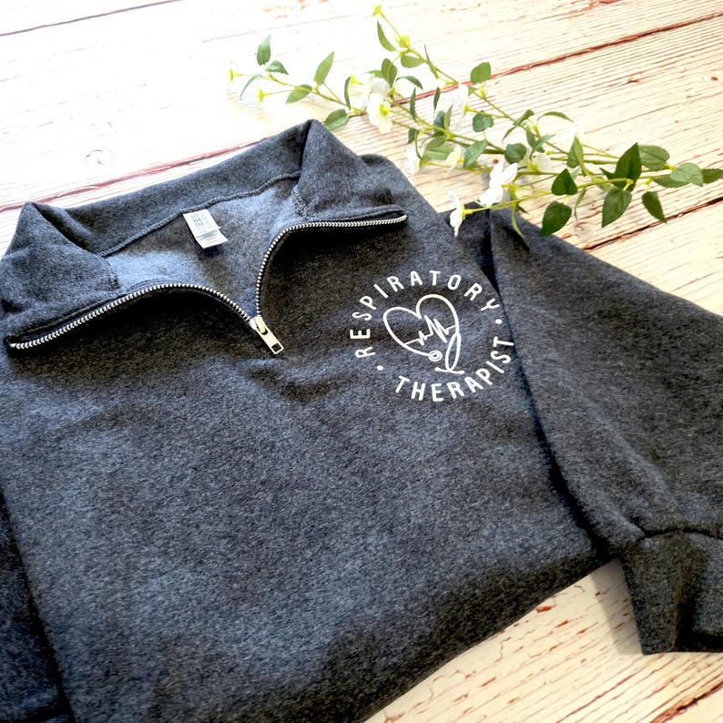 May include: A charcoal gray sweatshirt with a quarter-zip neckline. The sweatshirt features a white circular graphic with a heart, stethoscope, and the words "Respiratory Therapist". The garment is made of a soft material and has long sleeves.