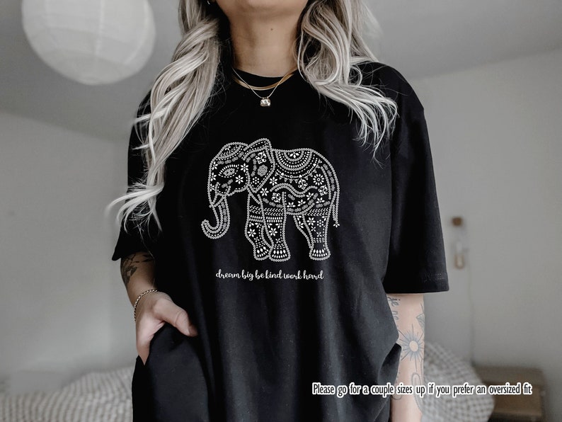 May include: A black t-shirt featuring a white graphic of an elephant decorated with intricate floral and geometric patterns. Below the elephant, the text reads "dream big be kind work hard". The bottom right corner includes the text "Please go for a couple sizes up if you prefer an oversized fit".