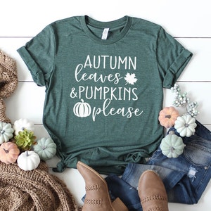 May include: A dark green t-shirt with the text "AUTUMN leaves & PUMPKINS please" in white. The shirt is styled with jeans, brown boots, and decorative pumpkins. A cozy brown scarf and fall-themed decor complete the autumn aesthetic.