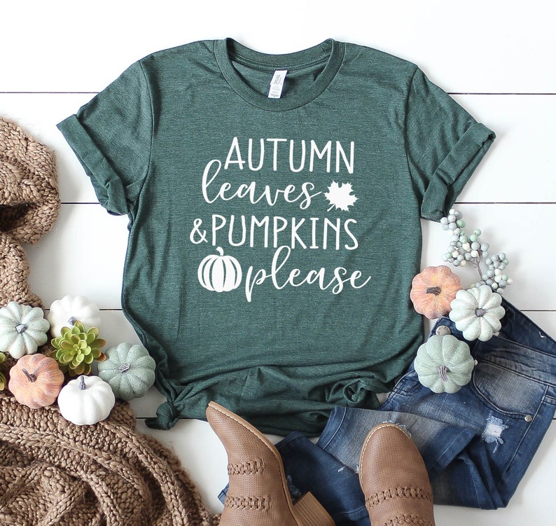 May include: A dark green t-shirt with the text "AUTUMN leaves & PUMPKINS please" in white. The shirt is styled with jeans, brown boots, and decorative pumpkins. A cozy brown scarf and fall-themed decor complete the autumn aesthetic.