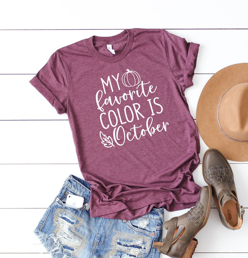 May include: A maroon t-shirt with the text "MY favorite COLOR IS October" in white, along with a pumpkin and leaf graphic. The shirt is paired with denim shorts, brown leather boots, and a tan hat.
