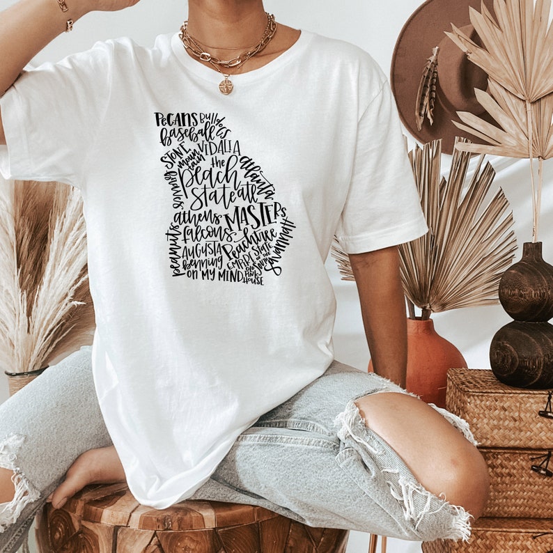 May include: White t-shirt featuring a black graphic of the state of Georgia filled with words like "Peaches," "Atlanta," and "Augusta." The shirt is a casual, crew neck style.