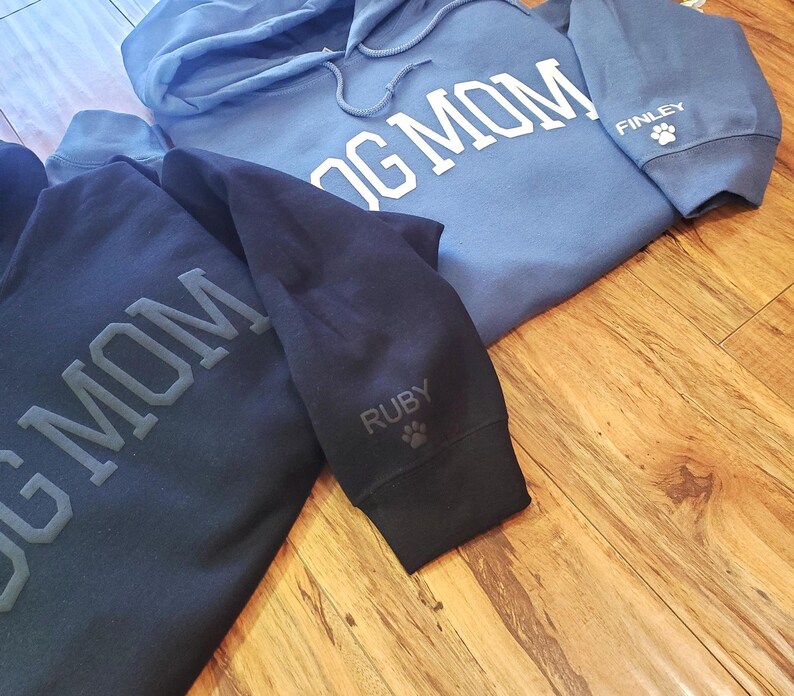 May include: Two hooded sweatshirts, one light blue and one navy blue, with the words "DOG MOM" printed across the chest. The light blue sweatshirt has "FINLEY" and a paw print on the sleeve. The navy blue sweatshirt has "RUBY" and a paw print on the sleeve.
