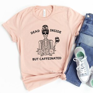 May include: A light pink t-shirt with the text "DEAD INSIDE BUT CAFFEINATED" and a black graphic of a skeleton wearing sunglasses and holding a coffee cup. The skeleton is surrounded by flowers.