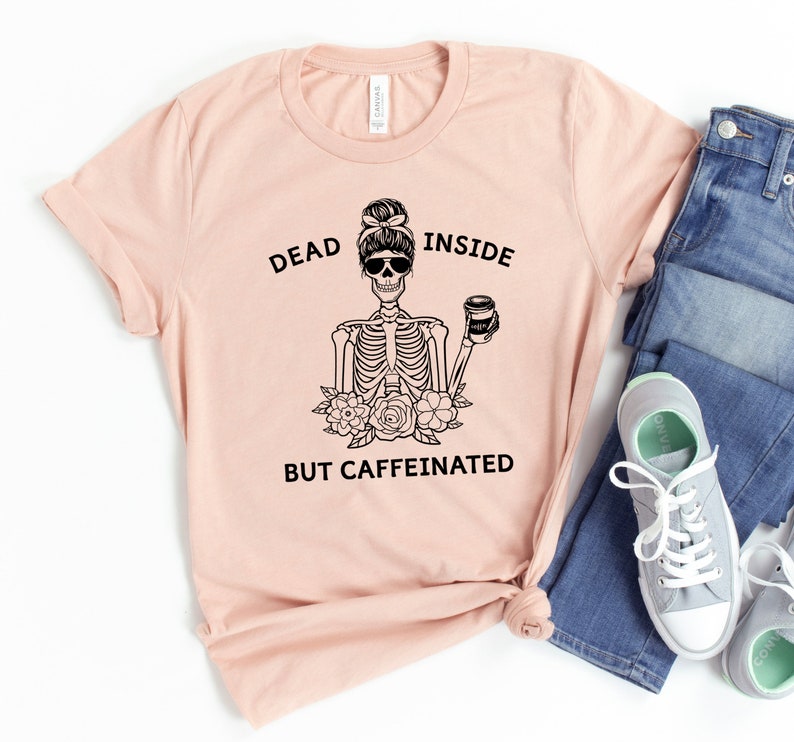 May include: A light pink t-shirt with the text "DEAD INSIDE BUT CAFFEINATED" and a black graphic of a skeleton wearing sunglasses and holding a coffee cup. The skeleton is surrounded by flowers.