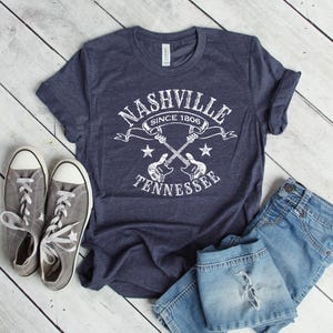 May include: Navy blue t-shirt with a white graphic that reads "Nashville Since 1806 Tennessee" with crossed guitars and stars. The shirt is paired with gray sneakers and blue jeans.