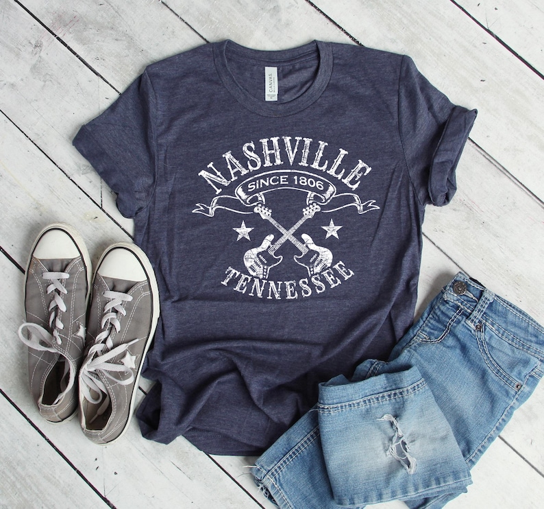 May include: Navy blue t-shirt with a white graphic that reads "Nashville Since 1806 Tennessee" with crossed guitars and stars. The shirt is paired with gray sneakers and blue jeans.