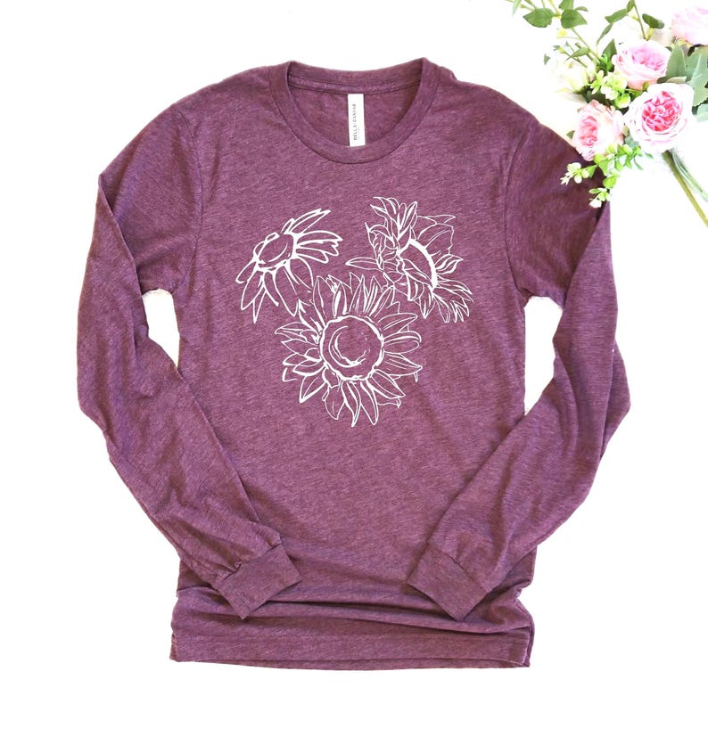 May include: A long-sleeved, burgundy-colored shirt featuring a white outline of three sunflowers. The floral design is centered on the front of the shirt. The shirt is made of a soft material and has a crew neck.