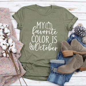 May include: Olive green t-shirt with the text "MY favorite COLOR IS October" in white. The shirt is styled with blue jeans, gray ankle boots, and a cozy sweater. A fall-themed design.