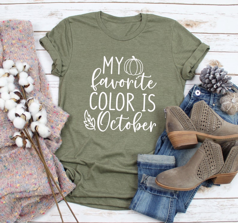 May include: Olive green t-shirt with the text "MY favorite COLOR IS October" in white. The shirt is styled with blue jeans, gray ankle boots, and a cozy sweater. A fall-themed design.