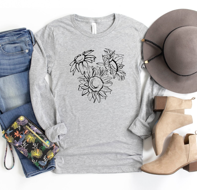 May include: A light gray long-sleeve shirt featuring a black line drawing of three sunflowers. The shirt is paired with blue jeans, a floral wallet, a gray hat, and tan ankle boots. The shirt is made of a soft, comfortable material.