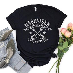 May include: Black t-shirt with a white distressed graphic that reads "Nashville Since 1806 Tennessee" with crossed guitars and stars. The shirt is styled with blue jeans, gray heels, and a bouquet of pink roses.