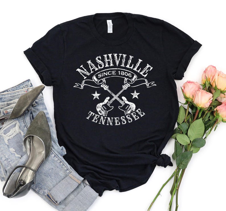 May include: Black t-shirt with a white distressed graphic that reads "Nashville Since 1806 Tennessee" with crossed guitars and stars. The shirt is styled with blue jeans, gray heels, and a bouquet of pink roses.