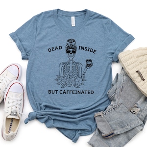 May include: A light blue t-shirt with the text "DEAD INSIDE BUT CAFFEINATED" and a black skeleton graphic wearing sunglasses and holding a coffee cup. The shirt is paired with white sneakers, jeans, and a cream-colored beanie.