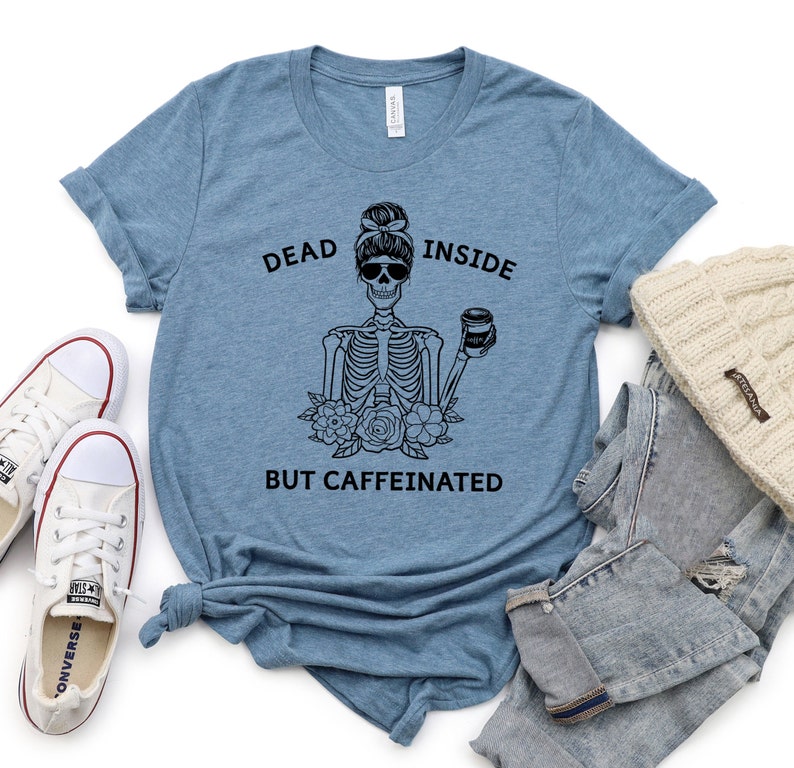 May include: A light blue t-shirt with the text "DEAD INSIDE BUT CAFFEINATED" and a black skeleton graphic wearing sunglasses and holding a coffee cup. The shirt is paired with white sneakers, jeans, and a cream-colored beanie.
