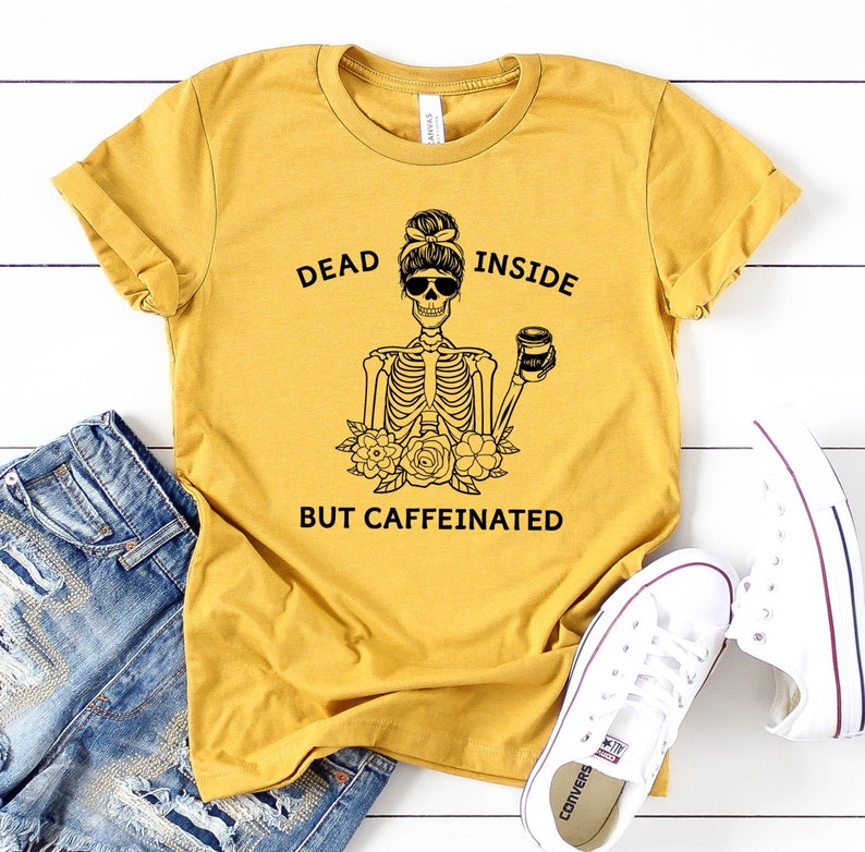 May include: Mustard yellow t-shirt with the text "DEAD INSIDE BUT CAFFEINATED" and a skeleton graphic. The skeleton is wearing sunglasses and holding a coffee cup. The shirt is paired with blue jeans and white sneakers.