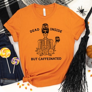 May include: Orange t-shirt with a black graphic of a skeleton wearing sunglasses and holding a coffee cup. The text reads "DEAD INSIDE BUT CAFFEINATED". The shirt is paired with blue jeans and Halloween decorations.