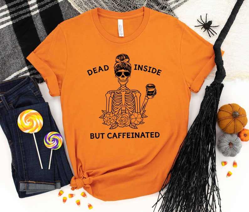 May include: Orange t-shirt with a black graphic of a skeleton wearing sunglasses and holding a coffee cup. The text reads "DEAD INSIDE BUT CAFFEINATED". The shirt is paired with blue jeans and Halloween decorations.