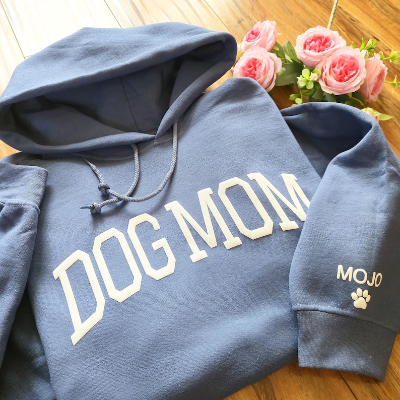 May include: A blue hoodie with the words "DOG MOM" in white block letters across the front. The sleeve features the word "MOJO" and a white paw print. The hoodie has a drawstring hood.