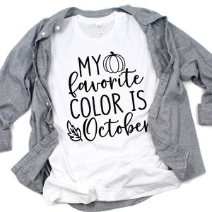 May include: White t-shirt with the text "MY favorite COLOR IS October" in black script, featuring a pumpkin and leaf graphic. A gray button-down shirt is draped over the t-shirt.