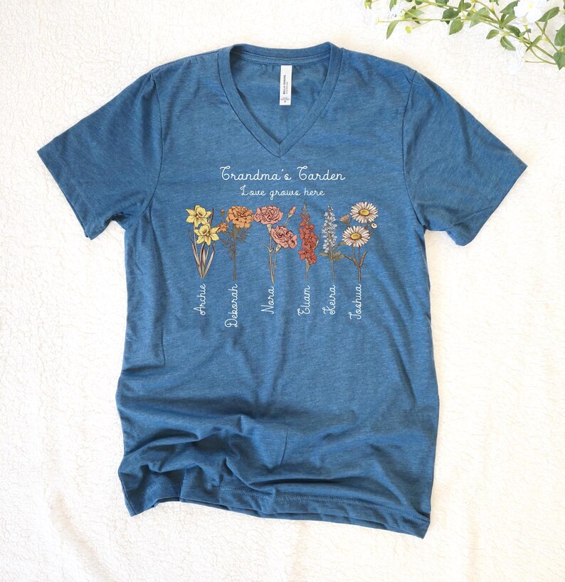 May include: Blue V-neck t-shirt with the text "Grandma's Garden Love grows here" and floral illustrations. The shirt features names like Archie, Deborah, and Joshua beneath the flowers. A casual, comfortable tee.
