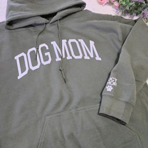May include: Olive green hooded sweatshirt with the text "DOG MOM" in white block letters across the chest. The sleeve features the text "UCY HOR" and a paw print. The sweatshirt has a front pocket and a drawstring hood.