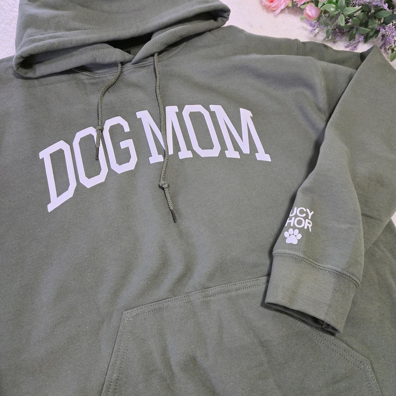 May include: Olive green hooded sweatshirt with the text "DOG MOM" in white block letters across the chest. The sleeve features the text "UCY HOR" and a paw print. The sweatshirt has a front pocket and a drawstring hood.