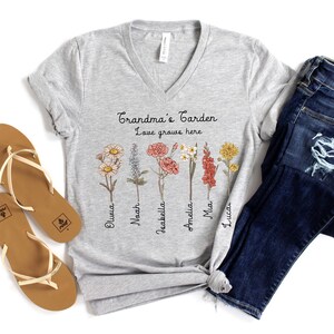 May include: A light gray V-neck t-shirt with the text "Grandma's Garden Love grows here" and floral illustrations. The shirt is paired with blue jeans and brown sandals.