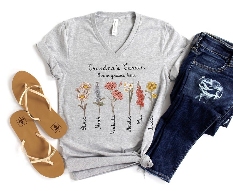 May include: A light gray V-neck t-shirt with the text "Grandma's Garden Love grows here" and floral illustrations. The shirt is paired with blue jeans and brown sandals.
