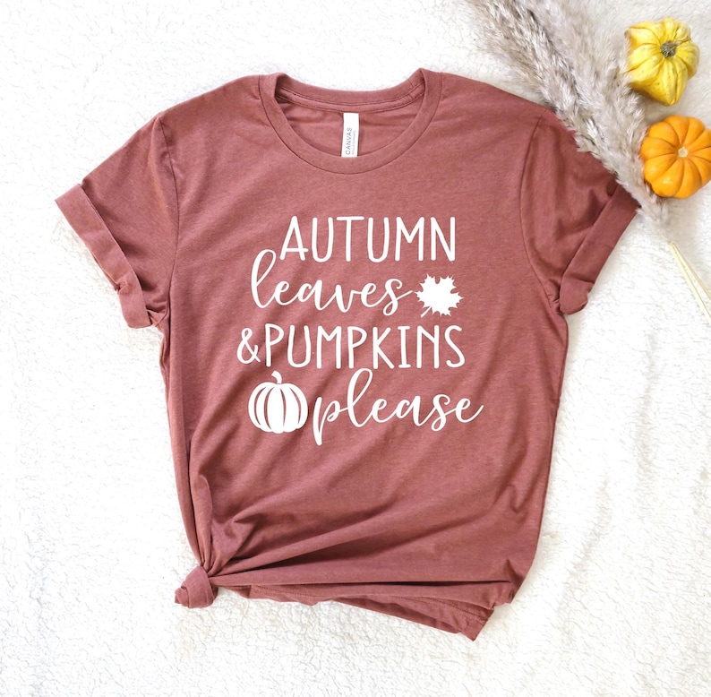 May include: A maroon t-shirt with rolled-up sleeves. The shirt features the text "AUTUMN leaves & PUMPKINS please" in white, along with a maple leaf and pumpkin graphic. The shirt is styled with fall-themed decorations.