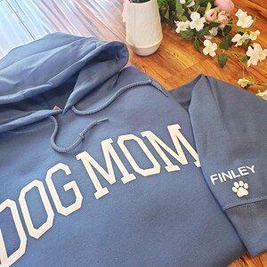 May include: A blue hoodie with the words "DOG MOM" in large white letters across the chest. The sleeve features the name "FINLEY" and a paw print. The hoodie has a drawstring hood.