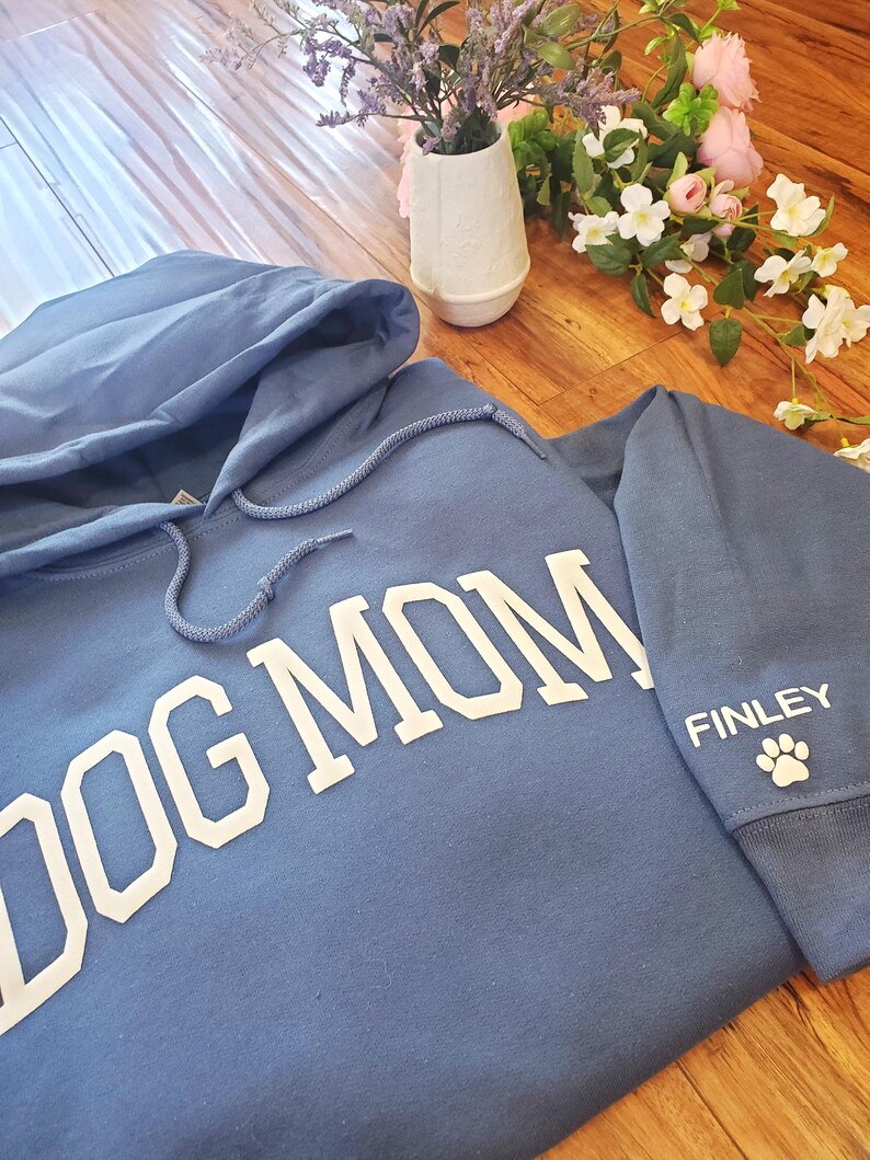 May include: A blue hoodie with the words "DOG MOM" in large white letters across the chest. The sleeve features the name "FINLEY" and a paw print. The hoodie has a drawstring hood.