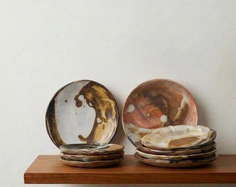 May include: A collection of ceramic plates and bowls featuring organic, irregular shapes and a hand-painted glaze finish. The pieces display a palette of earthy brown, cream, and terracotta tones with swirling, abstract patterns across the surfaces.
