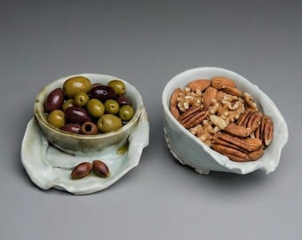 May include: Two light-colored ceramic bowls with organic, wavy edges. The left bowl contains a mix of green and dark purple olives. The right bowl is filled with a variety of shelled nuts, including almonds, walnuts, and pecan halves.