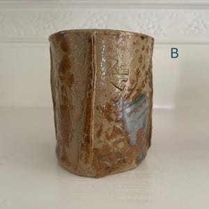 May include: A cylindrical ceramic mug with a textured, tan-colored surface featuring embossed geometric patterns and a vertical seam. A patch of blue glaze is visible on the side, and the letter B is printed in blue to the right of the mug.