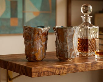 May include: Two handmade ceramic tumblers with an irregular, textured surface featuring a blend of earthy brown and muted grey-blue glaze. The cups have a thick, uneven rim and a cylindrical shape, positioned next to a glass decanter and a small glass.