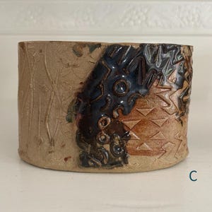 May include: A cylindrical ceramic vessel featuring a textured tan surface with embossed vertical lines and floral patterns. A prominent dark blue glazed section displays intricate carved geometric shapes and circular indentations. The letter C is visible.