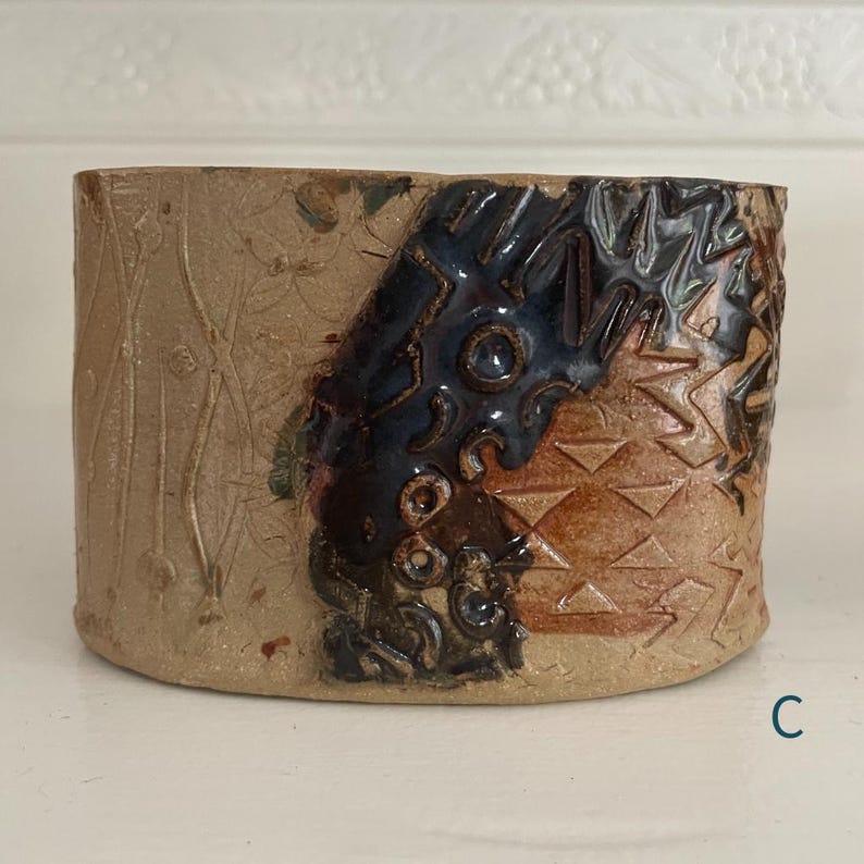 May include: A cylindrical ceramic vessel featuring a textured tan surface with embossed vertical lines and floral patterns. A prominent dark blue glazed section displays intricate carved geometric shapes and circular indentations. The letter C is visible.