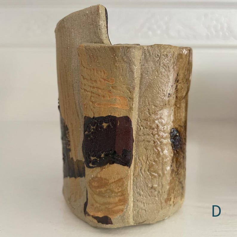 May include: A cylindrical ceramic vessel with a textured, beige, and tan surface featuring irregular vertical panels. The exterior displays dark brown, glossy, and matte patches with a visible letter D in the bottom right corner of the composition.