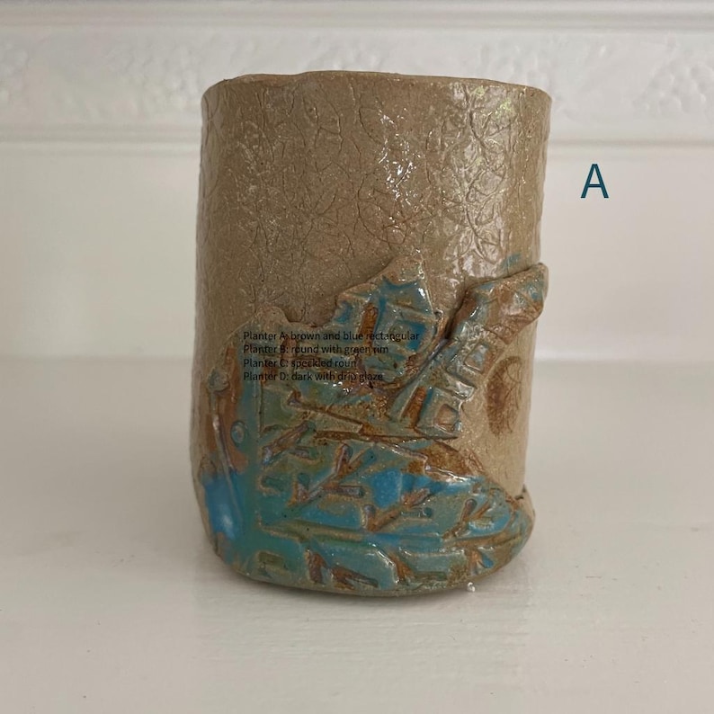 May include: A cylindrical ceramic planter with a textured tan surface featuring a raised, blue-glazed leaf design at the base. The letter A is printed in blue to the right of the object. Text on the planter reads: Planter A brown and blue rectangular.