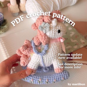May include: A white crocheted rocking horse toy with a pink mane, a light blue saddle, and a light blue base featuring small pink flower details. The text reads: PDF crochet pattern, Pattern update now available! See description for more info! by mariibun.